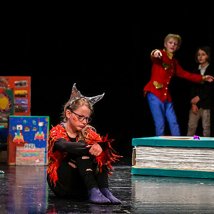 Ein Mädchen in Teufelskostüm sitzt mit einem frustrierten Gesichtsausdruck auf einer Bühne. Um sie herum liegen überdimensionierte Bücher. Im Hintergrund stehen zwei Kinder. Eines deutet auf das Mädchen.