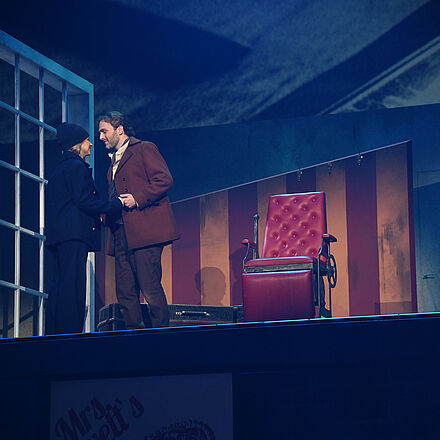 Eine Frau, in schwarzer Kleidung, und ein Mann stehen am Sessel. Sie schauen sich an