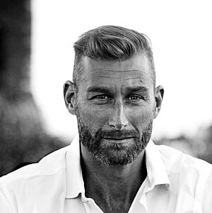 Schwarzweißes Portraitfoto von Florian Spiess. Er sitzt in ein weißes Hemd und eine dunkle Jeans gekleidet. Er sitzt. Sein linker Arm ist auf seinem linken Bein aufgestützt. Sein Haar ist an den Seiten kurz geschoren und oben gestylt. Er trägt einen Bart. Im Hintergrund ist eine Stadtsilhouette zu erahnen.