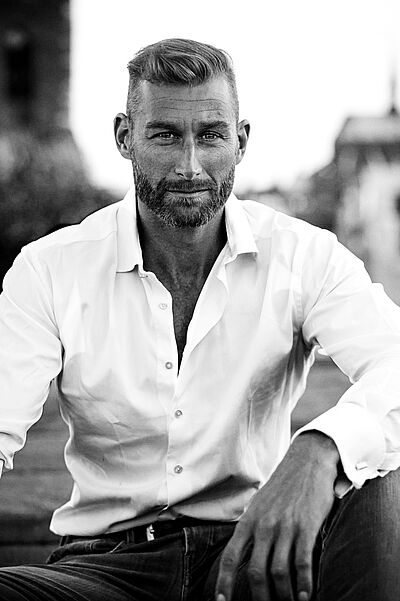 Schwarzweißes Portraitfoto von Florian Spiess. Er sitzt in ein weißes Hemd und eine dunkle Jeans gekleidet. Er sitzt. Sein linker Arm ist auf seinem linken Bein aufgestützt. Sein Haar ist an den Seiten kurz geschoren und oben gestylt. Er trägt einen Bart. Im Hintergrund ist eine Stadtsilhouette zu erahnen.