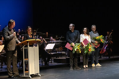 Drei Personen stehen mit Urkunden und Blumensträußen auf der Bühne vor dem Orchester. Links daneben spricht ein Herr am Pult.