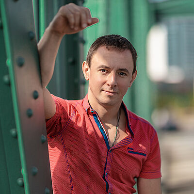 Portraitfoto von Volodymyr Fomenko. Er trägt ein rotes aufgeknöpftes T-Shirt mit blauen Akzenten. Sein rechter Arm lehnt an einer grünen Stahlstrebe.