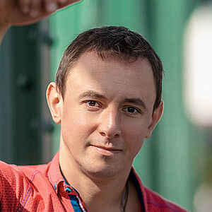 Portraitfoto von Volodymyr Fomenko. Er trägt ein rotes aufgeknöpftes T-Shirt mit blauen Akzenten. Sein rechter Arm lehnt an einer grünen Metallstrebe.