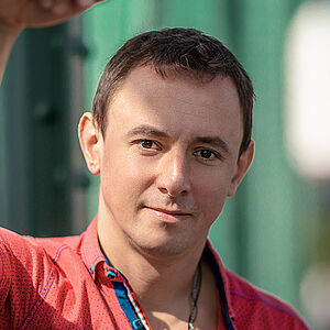 Portraitfoto von Volodymyr Fomenko. Er trägt ein rotes aufgeknöpftes T-Shirt mit blauen Akzenten. Sein rechter Arm lehnt an einer grünen Stahlstrebe.