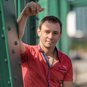 Portraitfoto von Volodymyr Fomenko. Er trägt ein rotes aufgeknöpftes T-Shirt mit blauen Akzenten. Sein rechter Arm lehnt an einer grünen Stahlstrebe.