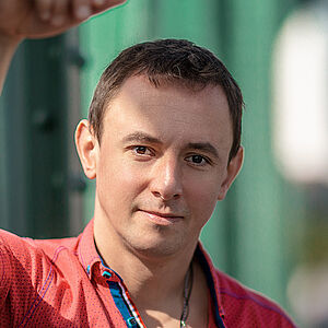 Portraitfoto von Volodymyr Fomenko. Er trägt ein rotes aufgeknöpftes T-Shirt mit blauen Akzenten. Sein rechter Arm lehnt an einer grünen Stahlstrebe.