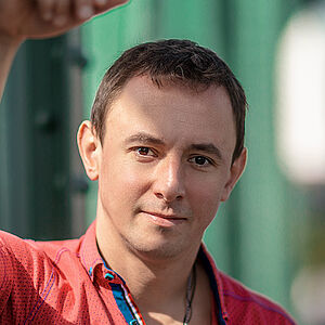 Portraitfoto von Volodymyr Fomenko. Er trägt ein rotes aufgeknöpftes T-Shirt mit blauen Akzenten. Sein rechter Arm lehnt an einer grünen Metallstrebe.