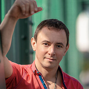 Portraitfoto von Volodymyr Fomenko. Er trägt ein rotes aufgeknöpftes T-Shirt mit blauen Akzenten. Sein rechter Arm lehnt an einer grünen Stahlstrebe.