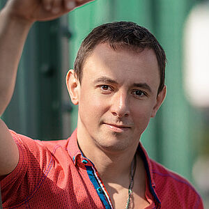 Portraitfoto von Volodymyr Fomenko. Er trägt ein rotes aufgeknöpftes T-Shirt mit blauen Akzenten. Sein rechter Arm lehnt an einer grünen Stahlstrebe.