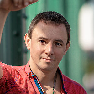 Portraitfoto von Volodymyr Fomenko. Er trägt ein rotes aufgeknöpftes T-Shirt mit blauen Akzenten. Sein rechter Arm lehnt an einer grünen Stahlstrebe.