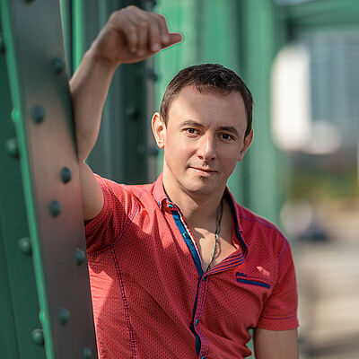 Portraitfoto von Volodymyr Fomenko. Er trägt ein rotes aufgeknöpftes T-Shirt mit blauen Akzenten. Sein rechter Arm lehnt an einer grünen Stahlstrebe.