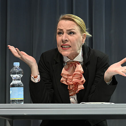 Eine Frau sitzt an einem Tisch. Sie hebt ihre Hände und Schultern an und blickt skeptisch nach vorne.