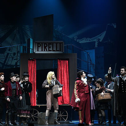 sehen ist eine große Personengruppe. Rechts im Vordergrund steht ein Mann mit einer Rasierklinge in der Hand. Links neben ihm steht ein Mann in roter Kleidung, der die Rasierklinge betrachtet. Weiter links, im Mittelpunkt des Vordergrunds, steht ein Mann mit langen, hellen Haaren, der eine braune Box in der Hand hält. Im Hintergrund der Personengruppe steht ein Händlerwagen mit roten Vorhängen und der Aufschrift «Pirelli».