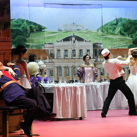Ein Mann sitzt vor einem Klavier und wird von einer Frau heruntergezogen. Im Hintergrund ist eine lange Tafel zu erkennen.