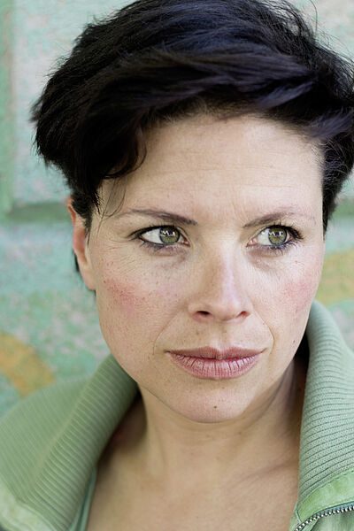 Portraitfoto von Sonja Elena Schroeder. Sie hat dunkles kurzes Haar und grüne Augen. Sie bliclt zur Seite und trägt ein hellgrünes Oberteil mit umgeschlagenem Kragen.