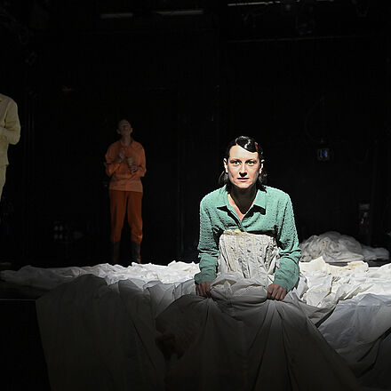 Die Frau im Kleid sitzt auf der Bühne. Im Hintergrund stehen drei Personen, sie essen Pommes.