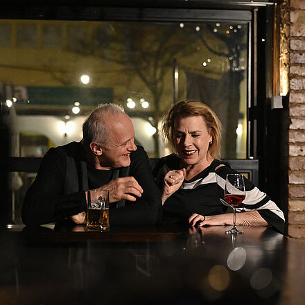 Die beiden Personen sitzen an der Bar und lächeln sich an. Vor ihnen stehen ein Glas Wein und ein Bier.