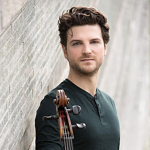 Portraitfoto von Gabriel Schwabe mit einem Cello. Er steht seitlich vor einer weißen Mauer. Er trägt ein dunkelgrüner Oberteil. Sein kurzes Haar ist braun und lockig. Er trägt einen Dreitagebart.