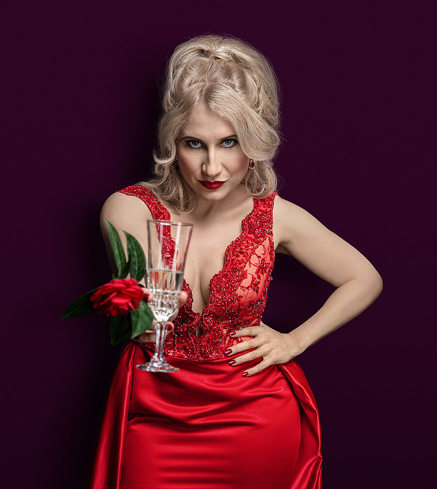 Eine Frau (Victoria Kunze) hat lange blonde lok´ckige Haare, die halb zurück gesteckt sind, während ihr zwei Strähnen ins GEsicht fallen. Die Frau trägt ein rotes Abendkleid und hält in iher nach vorn gestreckten Hand ein Champagnerglas und eine rote Kamelie. Der Hintergrund des Bildes ist bordeoux.
