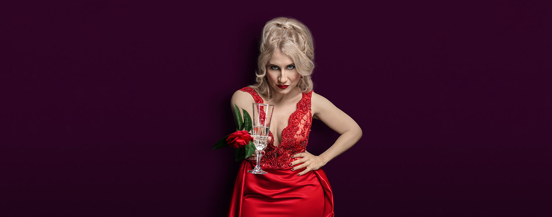 Eine Frau (Victoria Kunze) hat lange blonde lok´ckige Haare, die halb zurück gesteckt sind, während ihr zwei Strähnen ins GEsicht fallen. Die Frau trägt ein rotes Abendkleid und hält in iher nach vorn gestreckten Hand ein Champagnerglas und eine rote Kamelie. Der Hintergrund des Bildes ist bordeoux.
