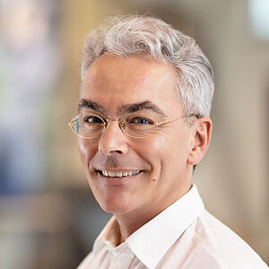 Portraitfoto von Hartmut Brüsch. Er trägt graues Haar, eine randlose Brille sowie ein weißes Hemd. Er lächelt mit geöffnetem Mund. 
