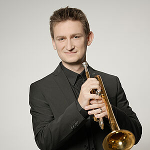Portraitfoto von Lennard Czakaj. Er hält seine Trompete vor dem Körper. Er ist in einen schwarzen Anzug gekleidet. Sein Haar ist an der Stirn hochgegelt. Der Hintergrund ist hellbeige.