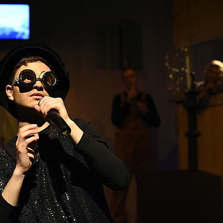 Ein Mann ist als Maulwurf verkleidet und trägt eine markante Brille. In seiner linken Hand hält er ein Mikrofon, in das er singt. Seine rechte Hand bewegt er zur Musik. Eine weitere Person befindet sich im Hintergrund und steht im Dämmerlicht der Bühnenbeleuchtung.