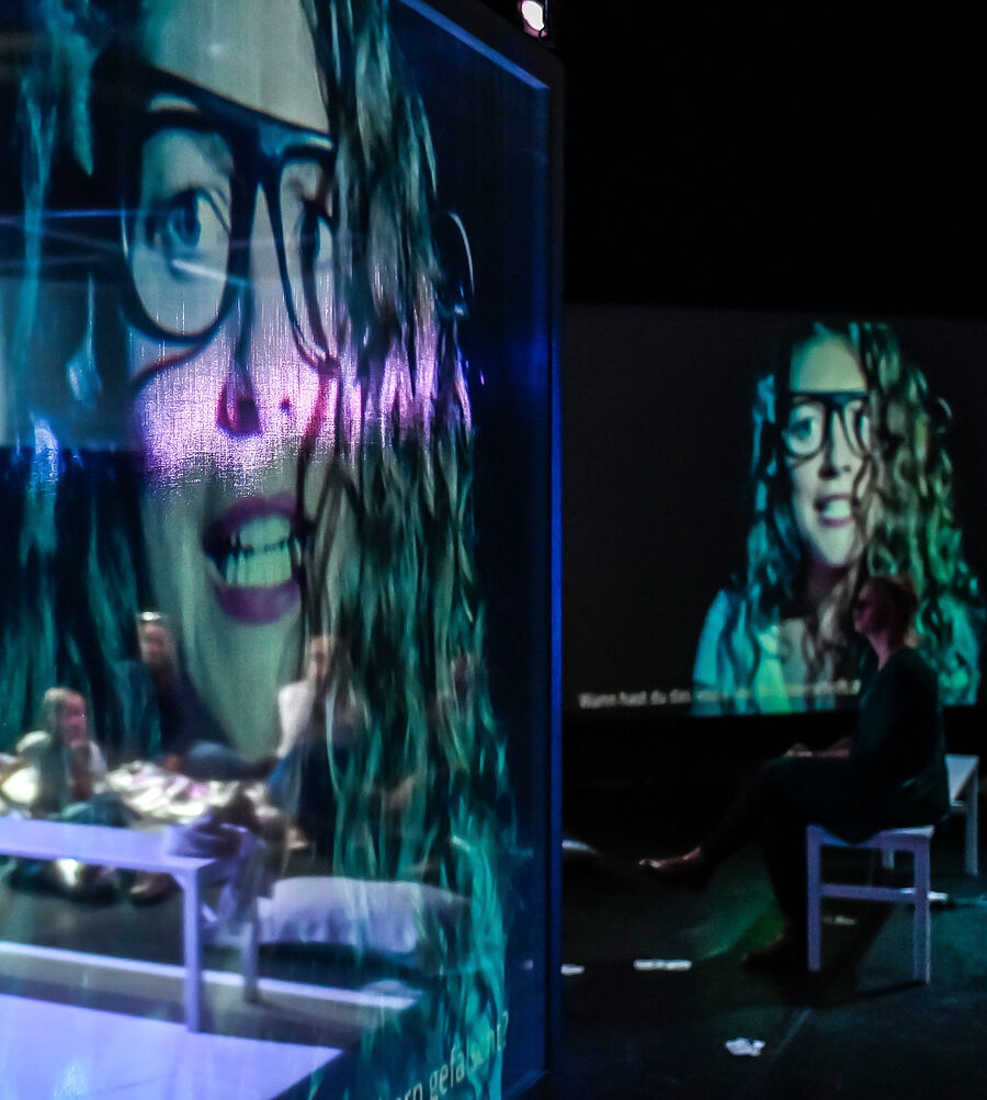Anna wird auf mehrere Leinwände projiziert. Sie hat lockiges langes Haar und trägt eine Brille. Sie spricht. Zwischen den Leinwänden sitzen Zuschauende auf Bänken, Sitzsäcken und am Boden und hören zu. Ihre Blicke sind auf die Leinwände gerichtet.