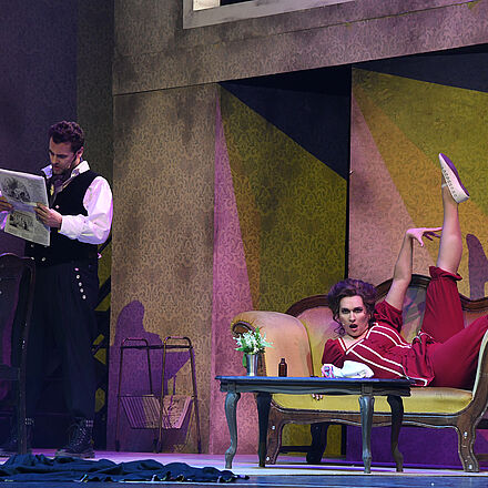 Links steht der Protagonist, er liest Zeitung. Auf dem Sofa liegt eine Frau.