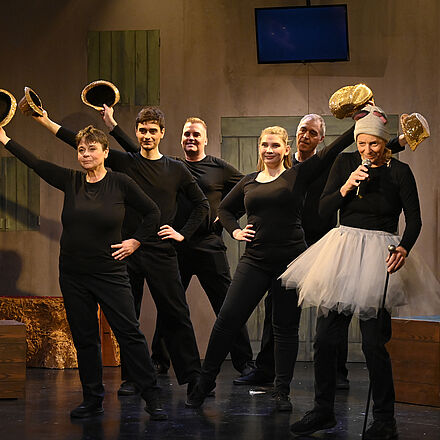 Fünf Personen halten fünf goldene Hüte in die Höhe. Ganz rechts steht eine sechste Person mit einem weißen Tutu, als Maus verkleidet und mit einem Mikrofon in ihrer linken Hand. 