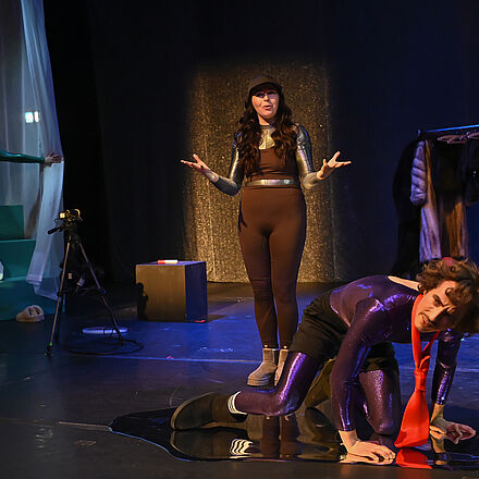 Die Person mit der Krawatte kniet auf dem Boden. Hinter ihr steht eine andere Person und schaut fragend. Im Hintergrund erkennt man Kleidung, eine Kamera und den Vorhang, aus dem die dritte Person in weißer Kleidung flüchtet.