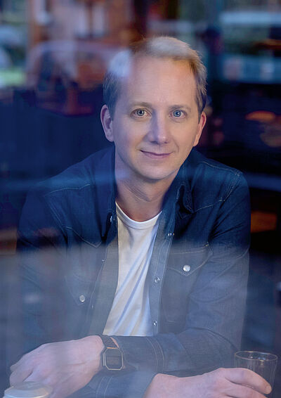 Portraitfoto von Dennis Gastmann durch eine Fensterscheibe.