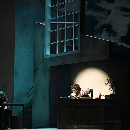 Links im Bild sitzt ein Mann an einem Tisch. Er schaut nach rechts. Rechts im Bild steht eine Frau hinter einem Tresen. Die Frau schaut nach links.