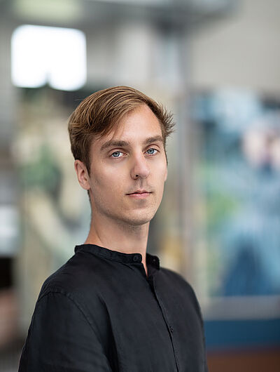 Portraitfoto von Torben Selk. Er steht seitlich. Er hat braunes Haar und trägt ein schwarzes Hemd ohne Kragen. Der Hintergrund ist verschwommen.