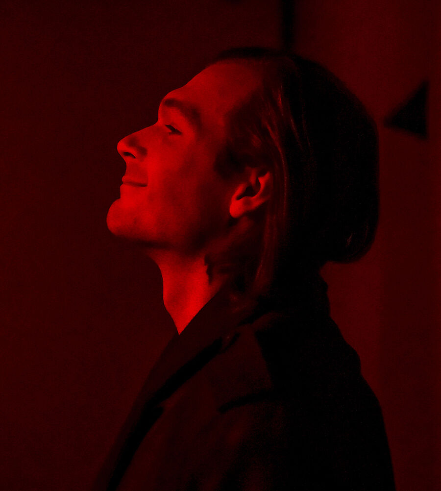 Demian steht seitlich. Er ist im Profil zu sehen. Seinen Kopf hat er in den Jacken gelegt. Er blickt zufrieden. Er herrscht eine rote Lichtstimmung. 