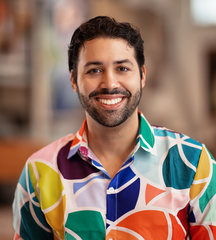 Portraitfoto von Mario El Fakih Hernández. Er trägt ein buntbedrucktes Hemd und lächelt mit geöffnetem Mund. Er trägt sein schwarzes Haar zurückgegelt und einen Dreitagebart.