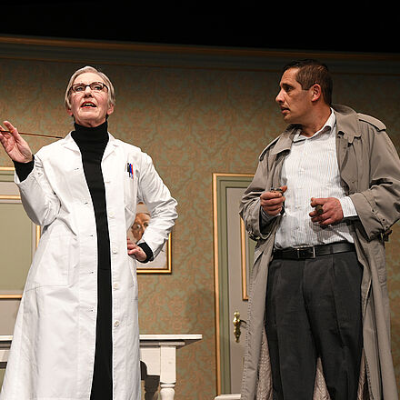 Links im Bild steht eine Frau mit einem Arztkittel und einer Zigarette in der Hand. Rechts steht ein Mann mit einem grauen Mantel.