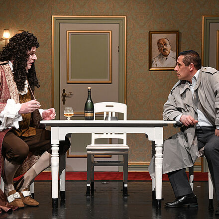 Im Mittelpunkt des Bildes steht ein Tisch. Links am Tisch sitzt ein Mann, der Kleidung und eine Perücke aus der Barock-Epoche trägt. Rechts am Tisch sitzt ein Mann, der einen grauen Mantel trägt und modern gekleidet ist. Im Hintergrund sieht man drei Türen.