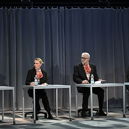 Zwei Frauen und zwei Männer sitzen jeweils nebeneinander an einem Tisch, in einer Reihe.