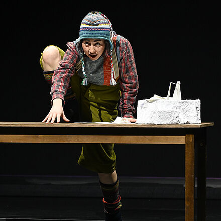 Eine Person klettert auf den Tisch. Ein kleines Podest aus Papier steht darauf 