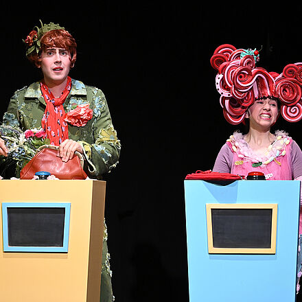 Zwei Personen stehen hinter jeweils einem Pult mit einem Knopf oben drauf.