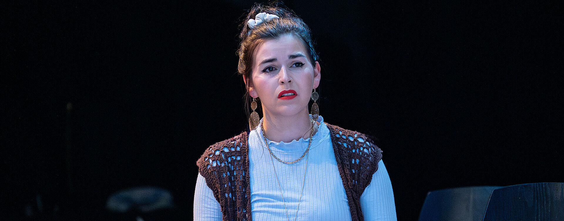 Eine junge Frau sitzt auf einem Stuhl, dem Publikum zugewandt. Sie scheint in traurigen Gedanken zu versinken. In ihren Händen hält sie ein kleines gelbes Buch.Sie trägt ein blaues Oberteil , eine braune Strickweste, einen roten Rock und schwarze Stiefel. Sie hat die Haare zu einem Dutt hochgesteckt.