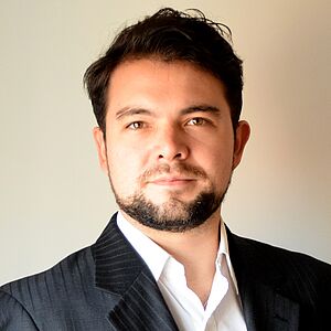 Portraitfoto von David Esteban. Er hat dunkelbraunes kurzes Haar und trägt einen Bart. Er ist in einen schwarzen Nadelstreifenanzug zu einem weißen Hemd gekleidet. Die Rückwand ist weiß.