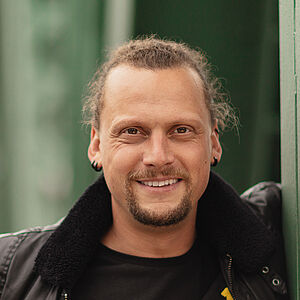 Portraitfoto von Henning Bäcker. Er lächelt. Sein Oberkörper lehnt an einem grünen Stahlgerüst. Sein linker Arm ist durch eine Öffnung der Streben gelegt. Er lächelt mit geöffnetem Mund. Er trägt eine schwarze Lederjacke mit weichem Kragen, Darunter ein schwarzes T-Short. Sein blondes Haar ist zu einem Zopf gebunden. Um seinen Mund trägt er Bart. In den Ohren sind schwarze Ohrringe.