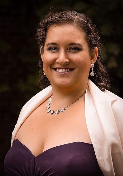 Portraitfoto von Mahina Gallinger. Sie lächelt mich geöffnetem Mund. Ihr dunkelbraunes Haar ist halb hochgesteckt. Sie trägt ein violettes trägerloses Kleid, dazu eine cremefarbene Stola und silbernen Schmuck.
