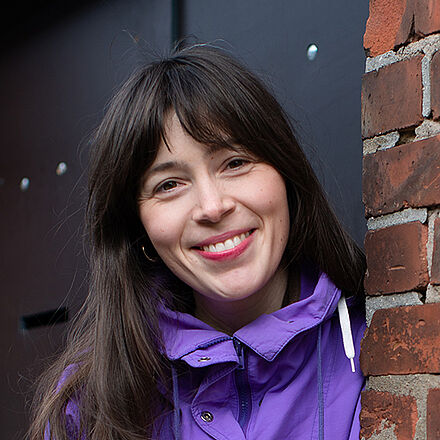 Portraitfoto von Meike Hoßbach in violetter Jacke. Sie hat dunkles Haar und lächelt in die Kamera. Sie lehnt an einer Ziegelwand.