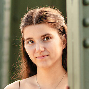 Portraitfoto von Zoe Irina Sauer Llano. Sie hat langes braunes Haar, das sie halbgeschlossen und mit Mittelscheitel trägt. Sie ist in ein ockerfarbenes Top gekleidet, dazu eine goldene Kette. Sie steht zwischen den Streben einer grünen Stahlbrücke und schaut hervor. Sie lächelt leicht. Der Hintergrund ist verschwommen.