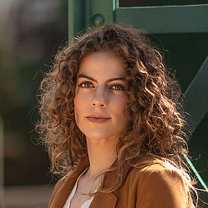 Portraitfoto von Alícia Navas Otero. Sie steht seitlich vor einer grünen Stahlstrebe. Ihr lockiges braunes Haar legt sich über ihre Schultern. Sie trägt einen camelfarbenen Blazer, darunter eine weiße Spitzenbluse. 
