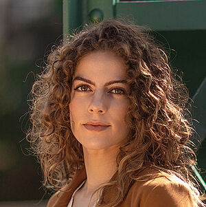 Portraitfoto von Alícia Navas Otero. Sie steht seitlich vor einer grünen Stahlstrebe. Ihr lockiges braunes Haar legt sich über ihre Schultern. Sie trägt einen camelfarbenen Blazer, darunter eine weiße Spitzenbluse. 