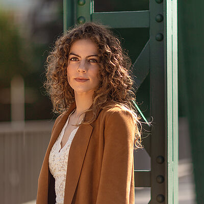 Portraitfoto von Alícia Navas Otero. Sie steht seitlich vor einer grünen Stahlstrebe. Ihr lockiges braunes Haar legt sich über ihre Schultern. Sie trägt einen camelfarbenen Blazer, darunter eine weiße Spitzenbluse.
